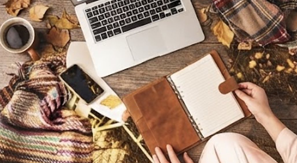 A laptop, a notebook and a coffee on a wooden table with autumn leaves